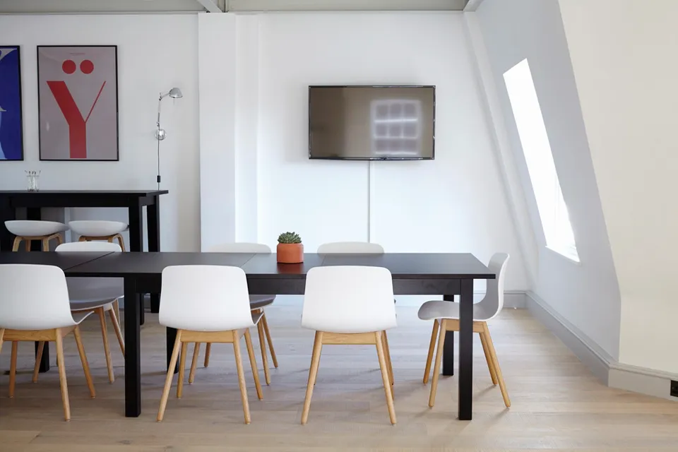 Minimal meeting room with a dark table, white chairs, a wall-mounted TV, small cactus and framed poster, lit by a slanted skylight