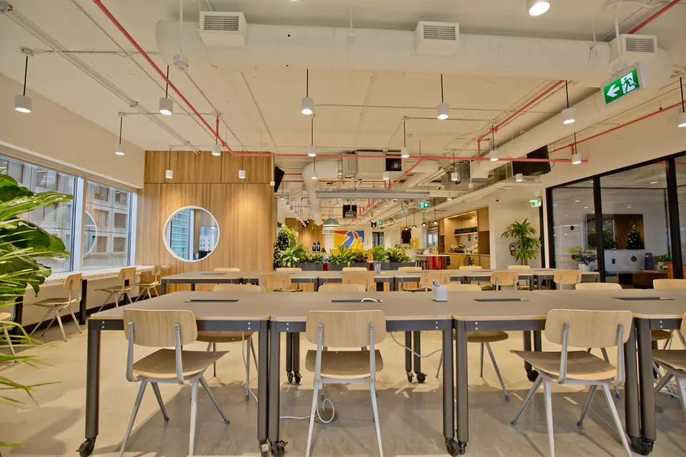 Open hot-desk area with rows of long tables and light wood chairs, exposed ducts and red pipes, pendant lights, plants and big windows
